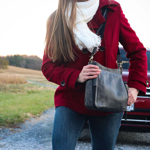 woman holding a Gray distressed leather crossbody concealed carry bag with RFID-blocking card storage, vertical top-draw gun access, supports left/right-handed draw, secure fit for compact handguns, anti-slash strap included