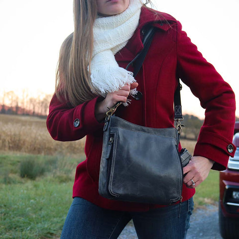 woman holding a Gray distressed leather crossbody concealed carry bag tightly and grabbing a pistol out of it  with RFID-blocking card storage, vertical top-draw gun access, supports left/right-handed draw, secure fit for compact handguns, anti-slash strap included